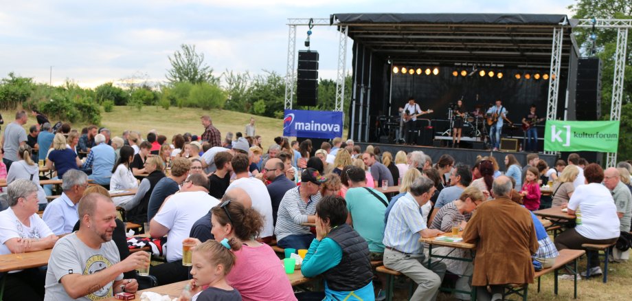 Viele Menschen an Biertischen vor einer großen Bühne.