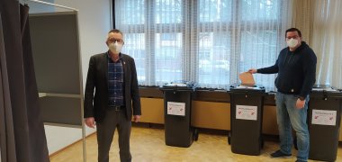 Jirasek und Sander zeigen im Briefwahlbüro Abstimmungskabine und Wahlurne in Mülltonnen-Form.