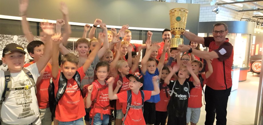 Der Bürgermeister und die Ferienspielkinder halten einen Pokal im Eintrachtmuseum in die Luft!