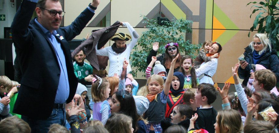 Der Bürgermeister im Rathausfoyer umgeben von verkleideten Kitakindern animiert mit erhobenen Händen zum lauten "Helau"-Ruf.