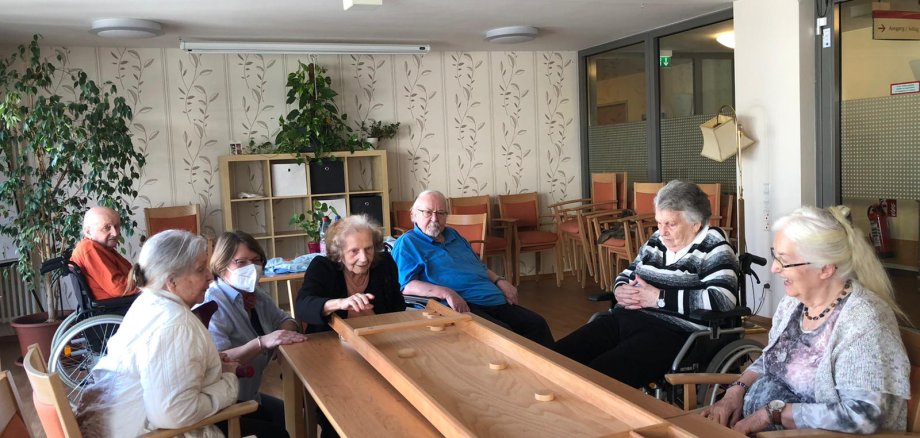 Senioren sitzen um ein Shuffleboard herum. Eine Dame schiebt den Puk.