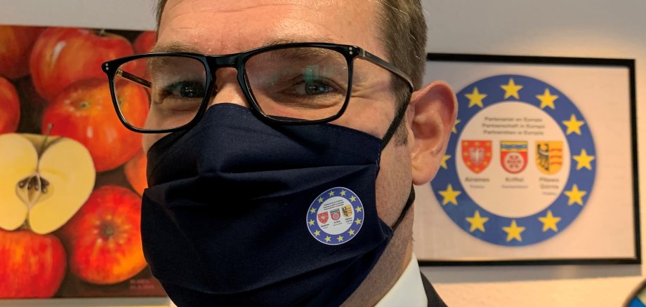 Bürgermeister Seitz trägt eine blaue Maske mit dem Logo der Partnerschaft Kriftel - Airaines - Pilawa Gorna.