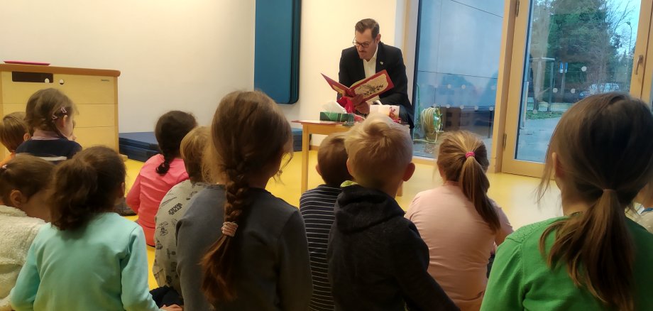 Um den Bürgermeister mit Buch in der Hand sitzen viele Kinder herum.