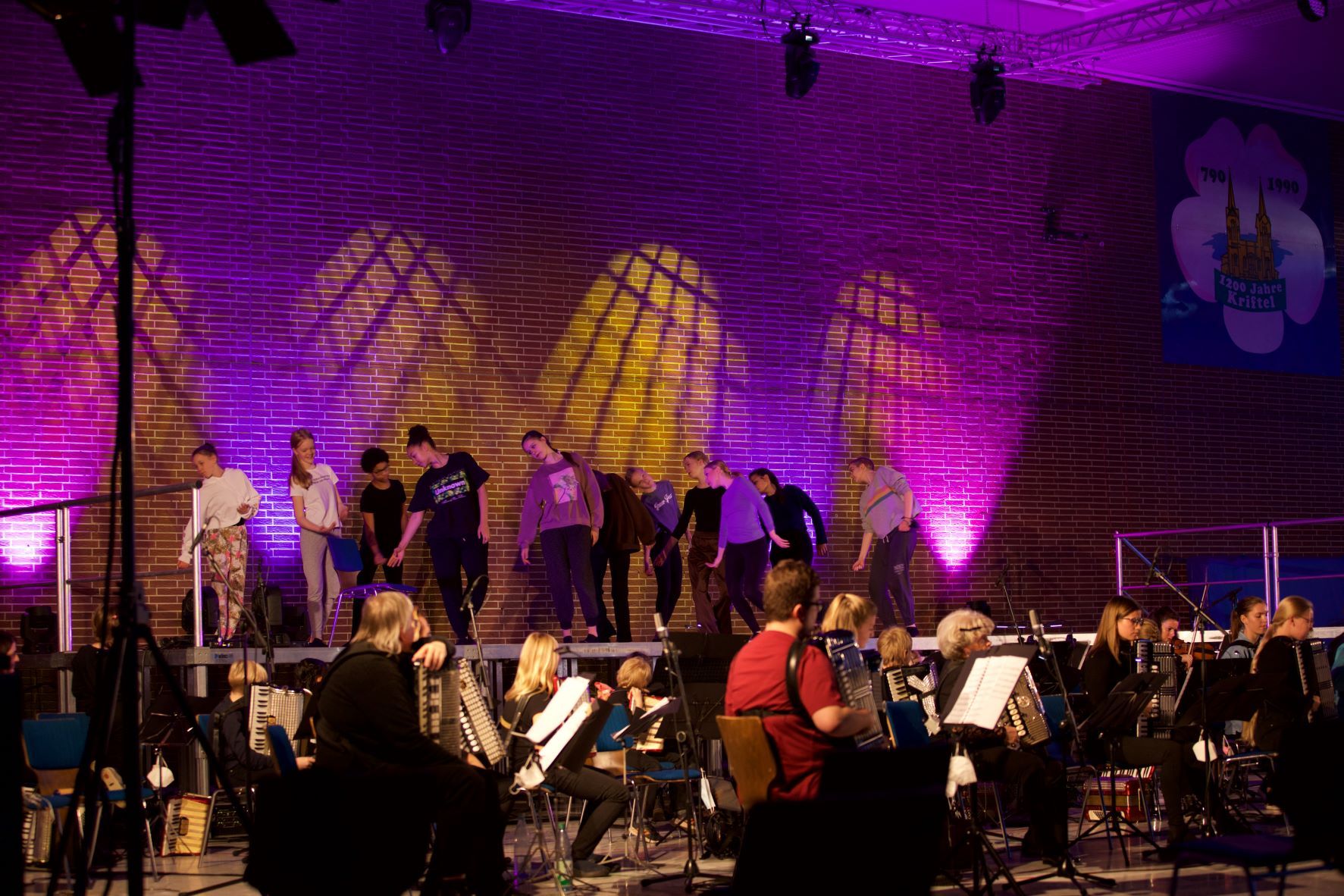 Tänzer auf der Bühne, davor Akkordeonspieler. Alles Lila beleuchtet.