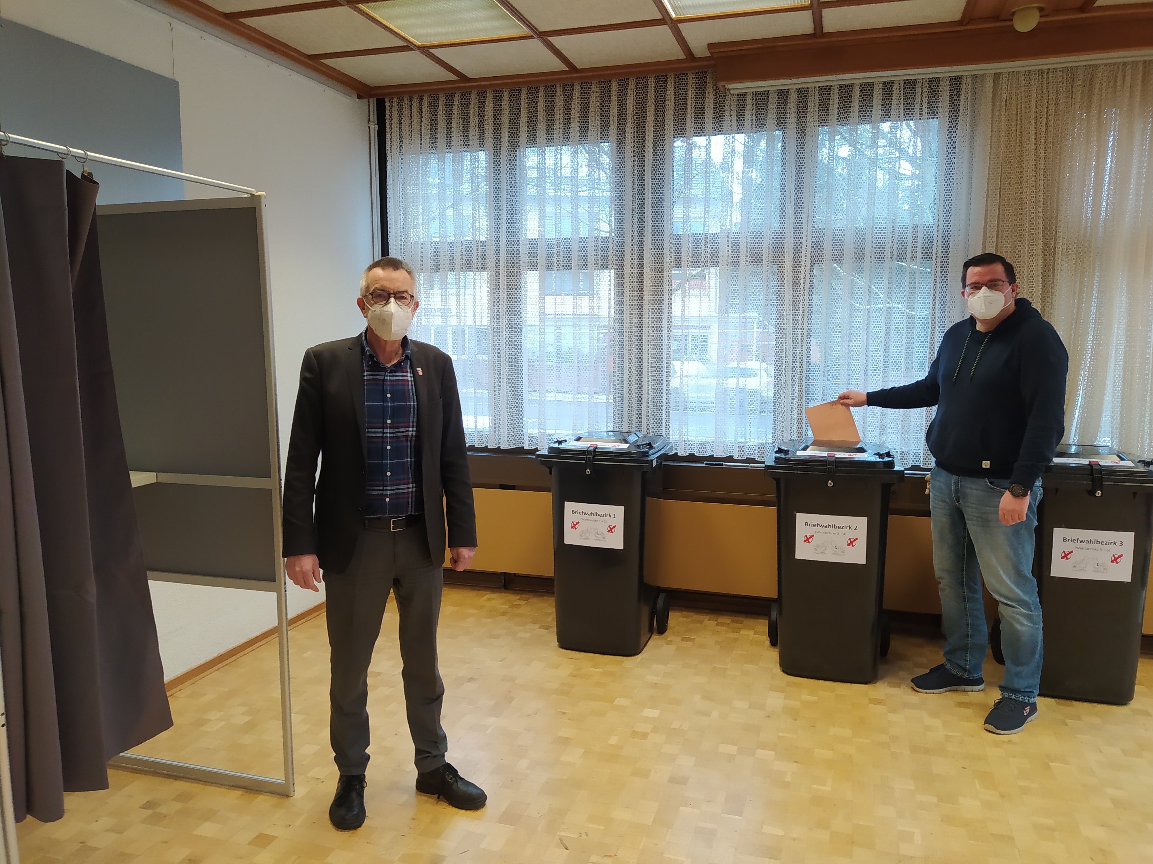 Jirasek und Sander im Wahlbüro vor Wahlurnen und Wahlkabine.