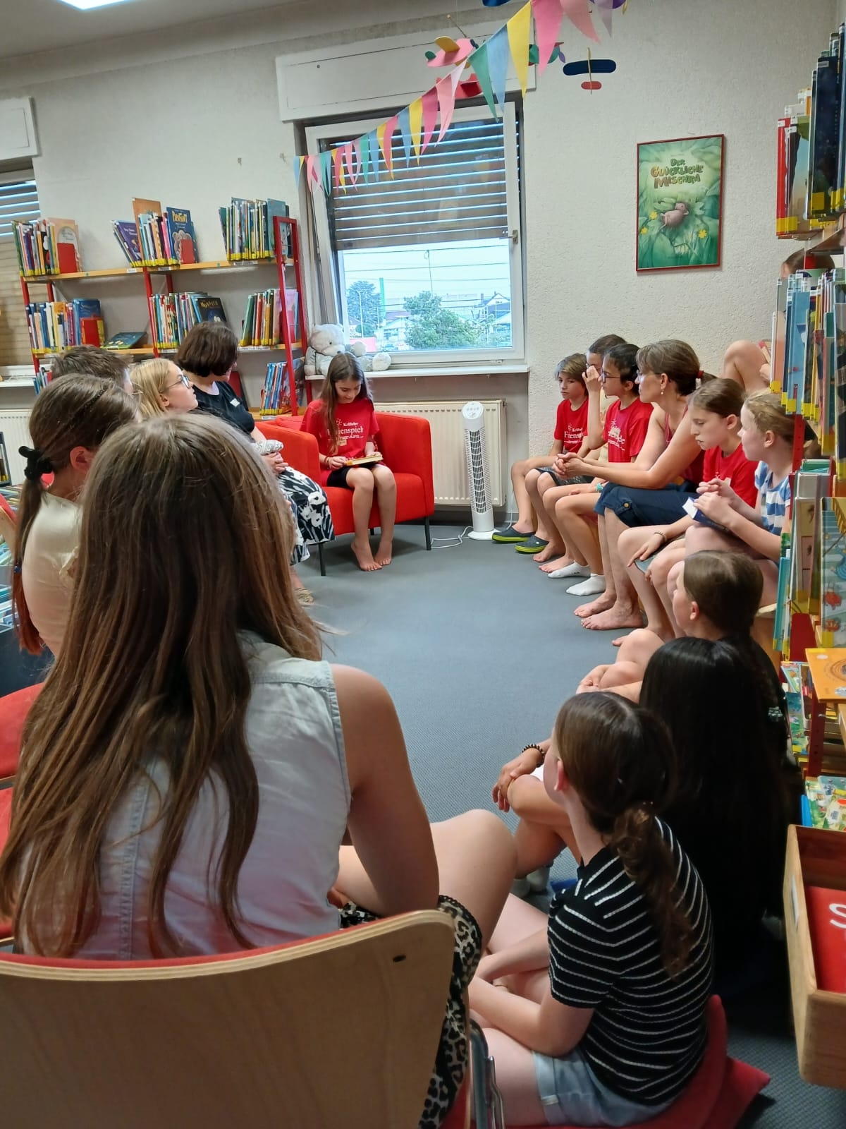 Kinder sitzen im Kreis zwischen Bücherregalen.