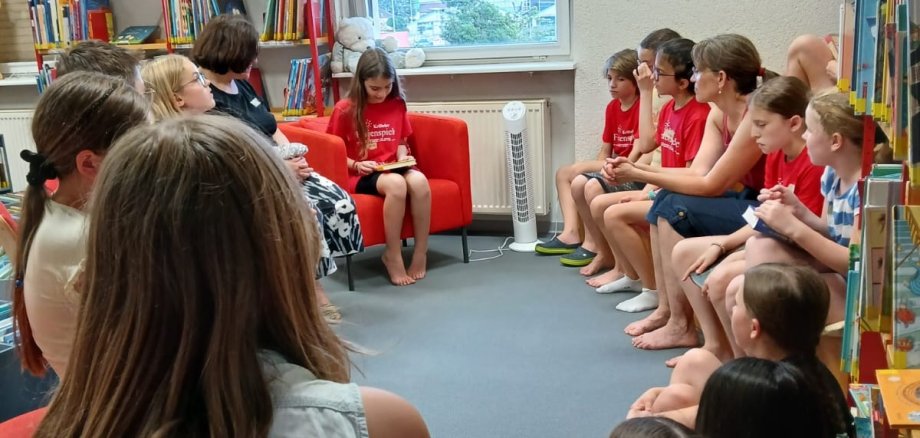 Kinder sitzen im Kreis zwischen Bücherregalen.