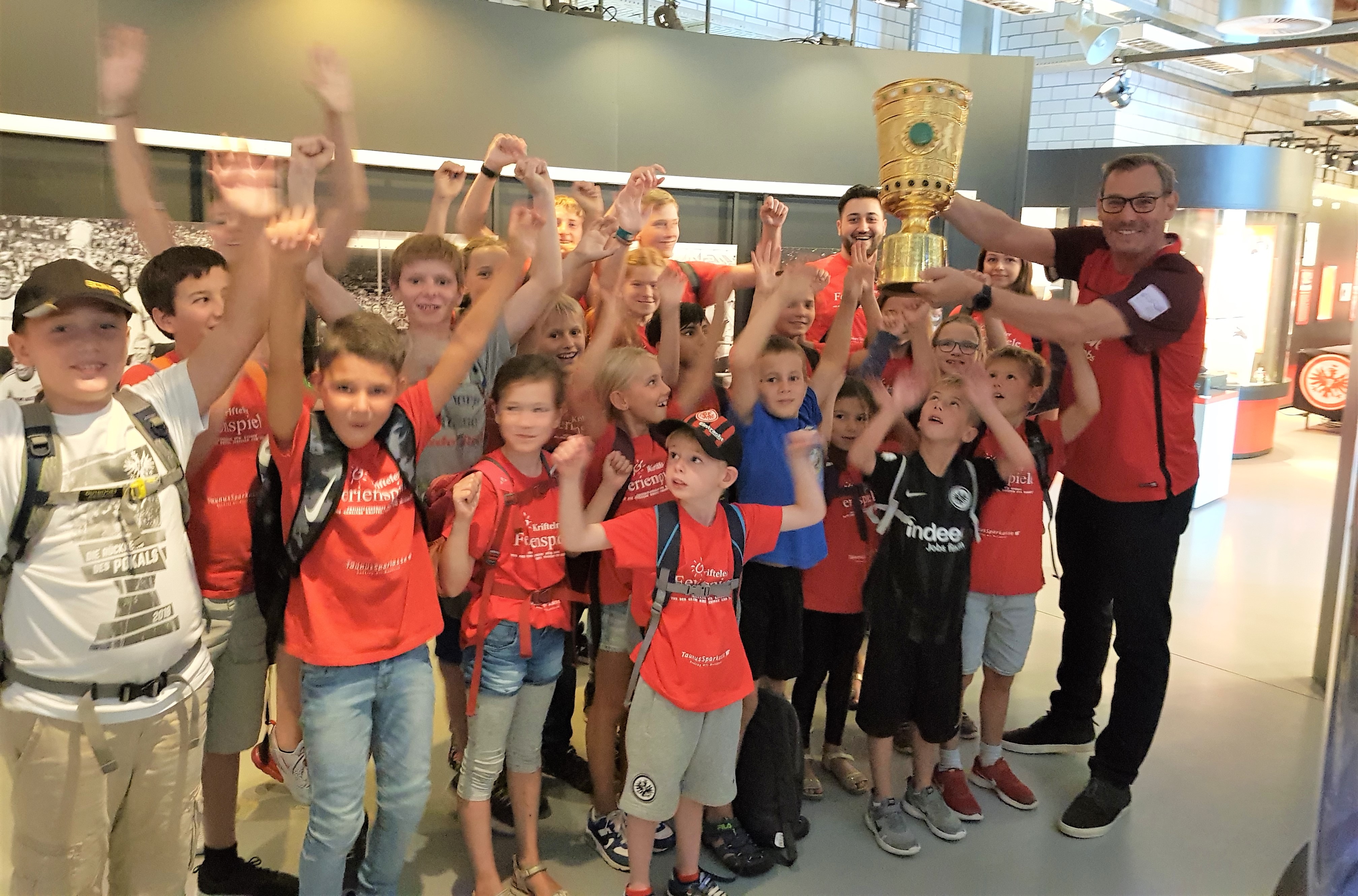 Der Bürgermeister und die Ferienspielkinder halten einen Pokal im Eintrachtmuseum in die Luft!