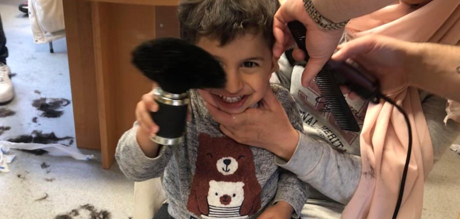 Ein Kind mit Frisörpinsel bekommt auf dem Schoss der Mutter die Haare geschnitten.
