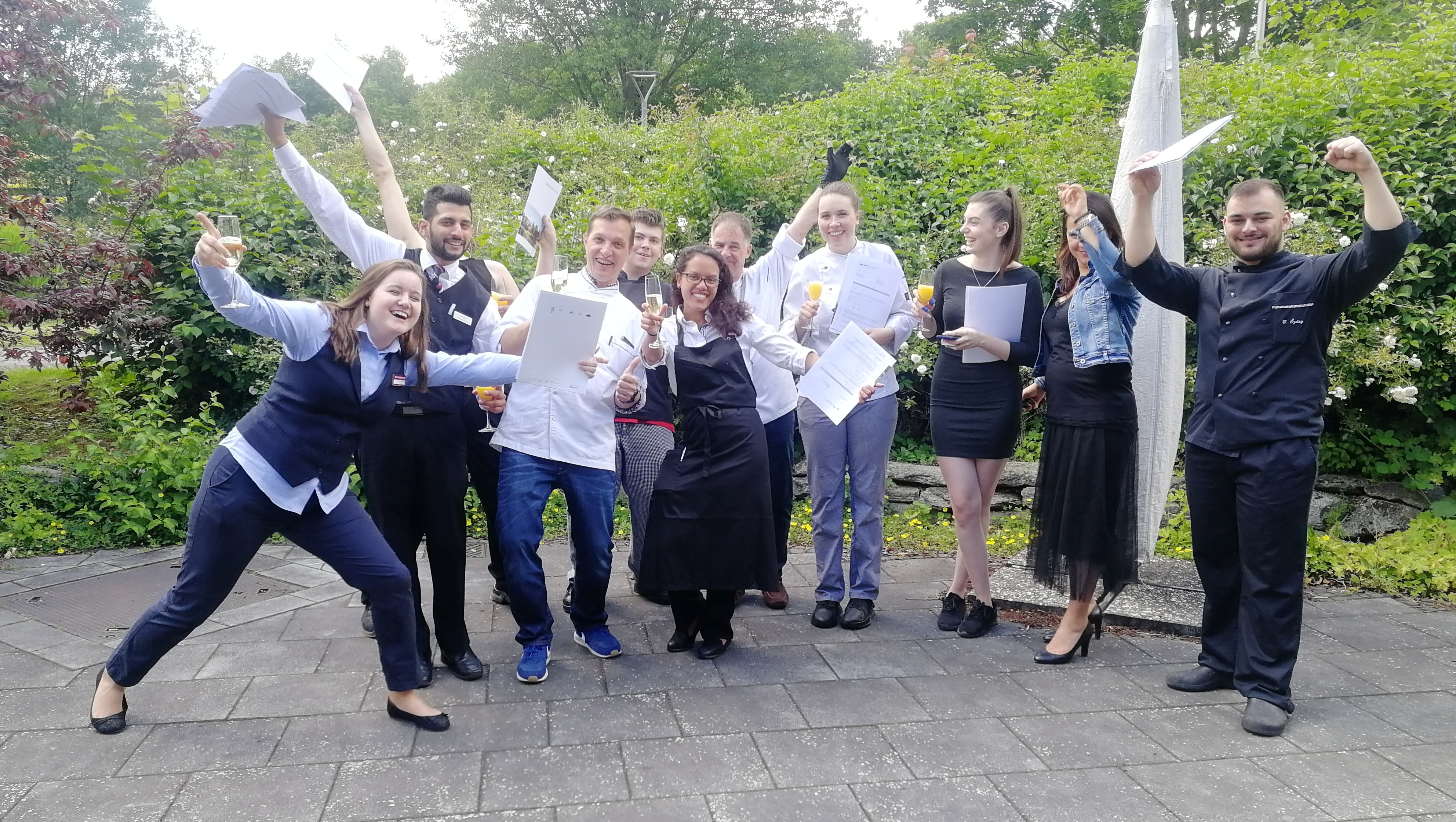 Freudige junge Menschen in Kochoutfit halten ihre Zeugnisse hoch.