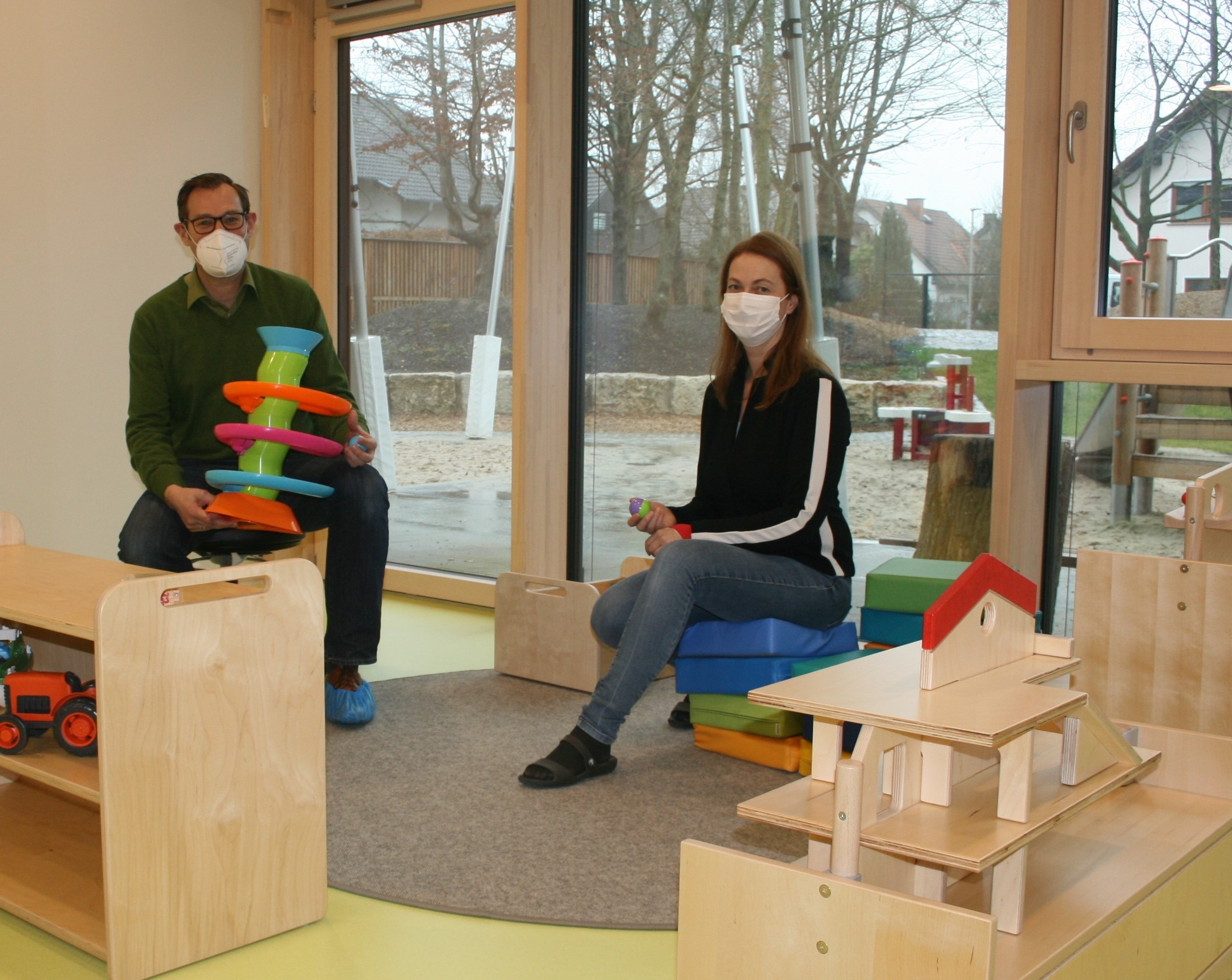 Bürgermeister Seitz und Kitaleiterin Bernstein mit Masken und Spielzeug im Gruppenraum für die Kleinsten.