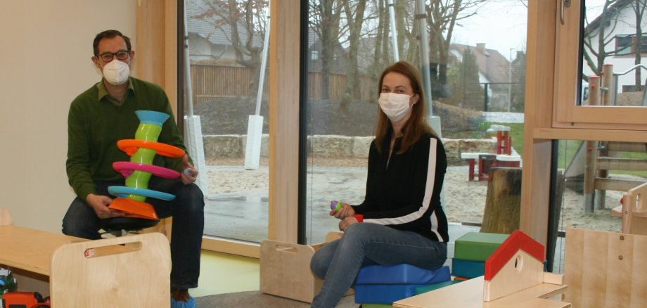 Bürgermeister Seitz und Kitaleiterin Bernstein mit Masken und Spielzeug im Gruppenraum für die Kleinsten.