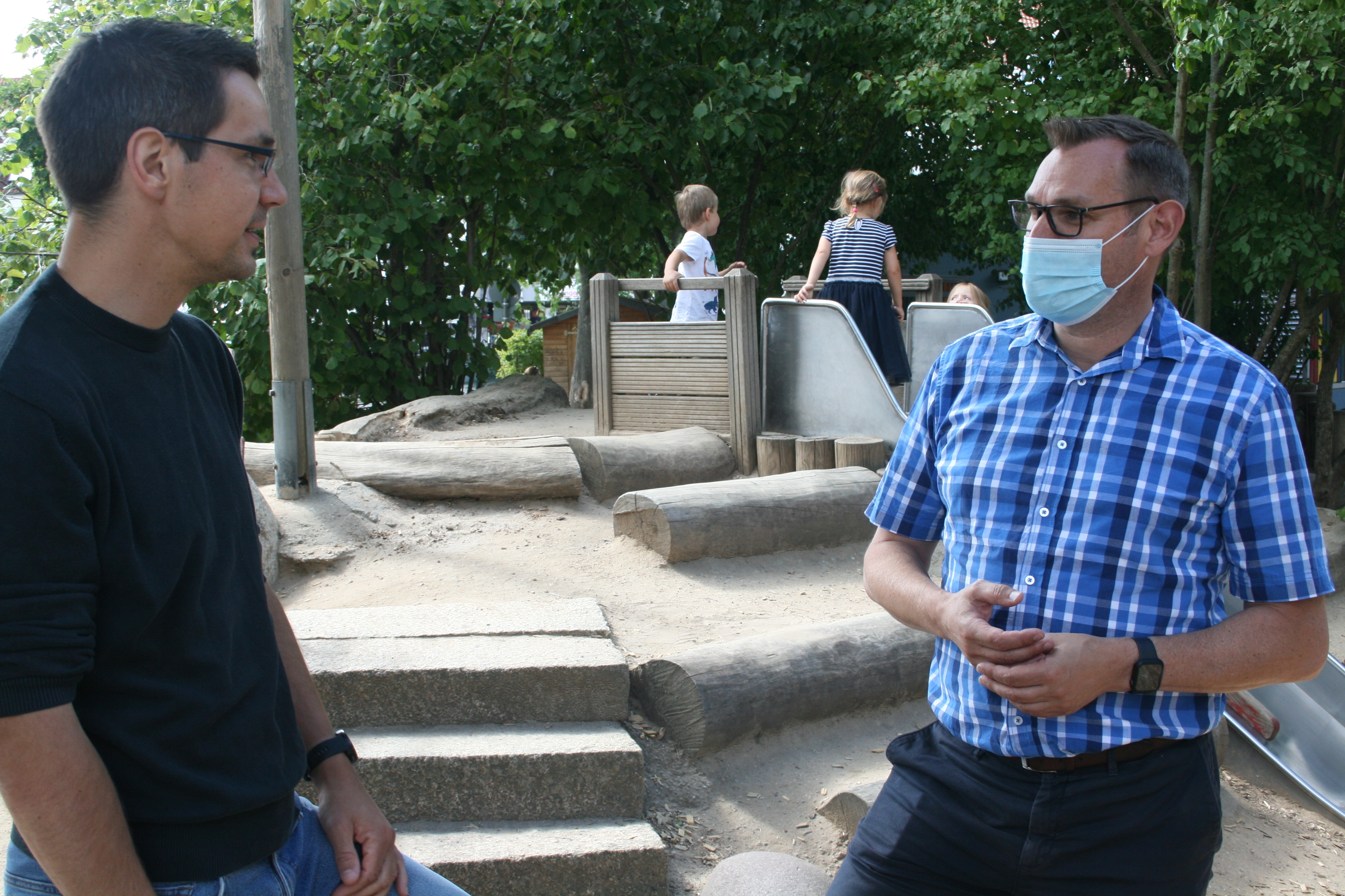 Bürgermeister Seitz im Gespräch mit einem Erzieher der Kita Vogelnest im Außenbereich. Im Hintergrund spielende Kinder.