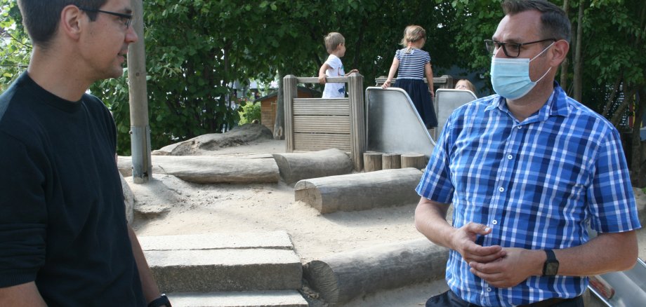 Bürgermeister Seitz im Gespräch mit einem Erzieher der Kita Vogelnest im Außenbereich. Im Hintergrund spielende Kinder.