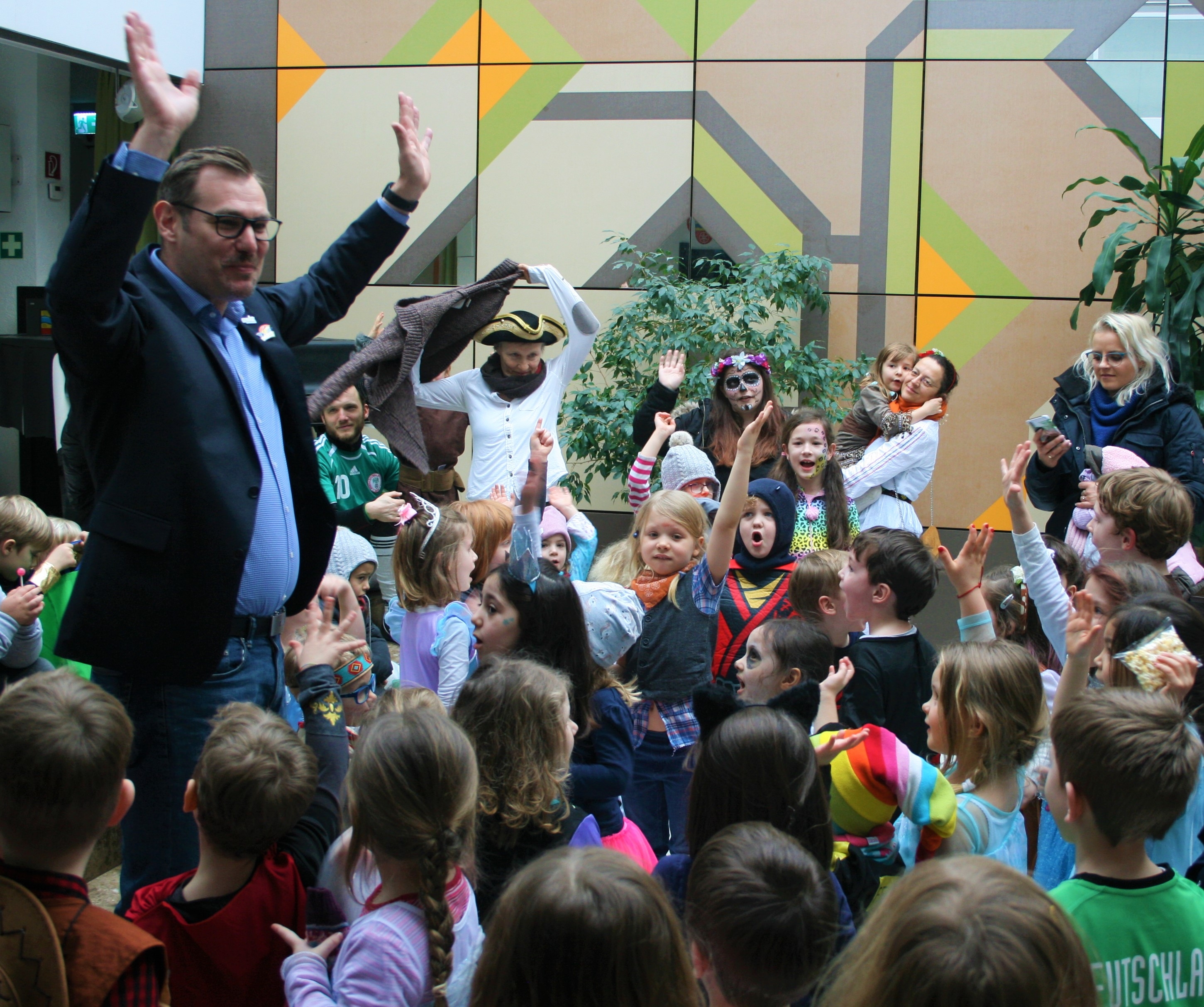 Der Bürgermeister im Rathausfoyer umgeben von verkleideten Kitakindern animiert mit erhobenen Händen zum lauten "Helau"-Ruf.