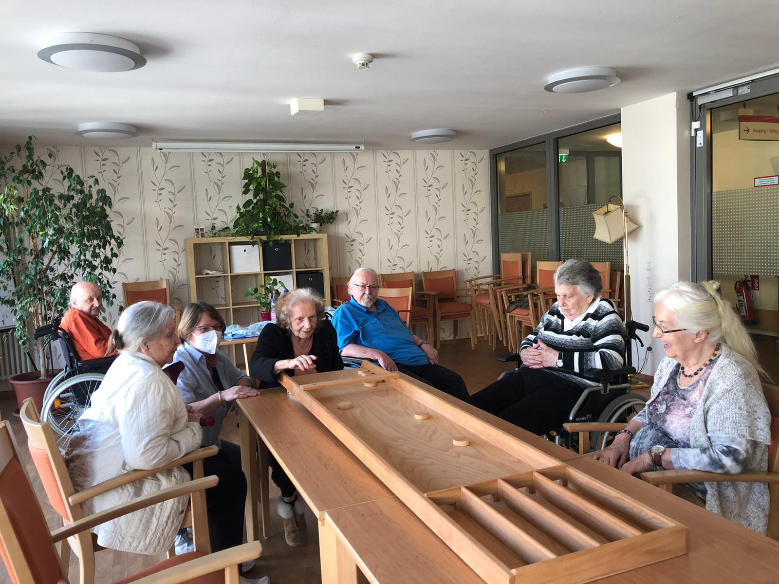 Senioren sitzen um ein Shuffleboard herum. Eine Dame schiebt den Puk.