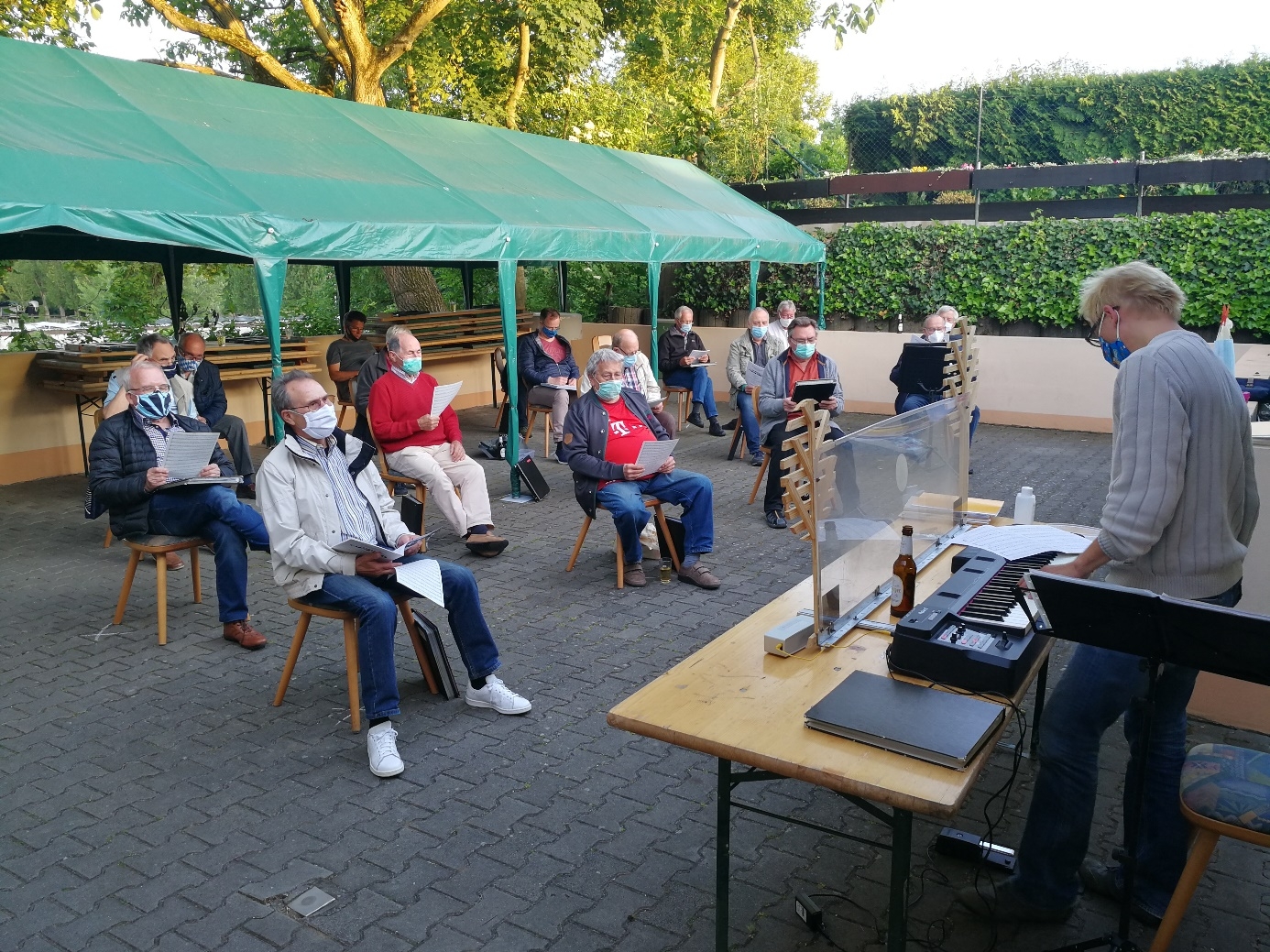 Die Mitglieder des Männerchors sitzen mit Masken auf Stühlen und proben im Freien mit viel Abstand.