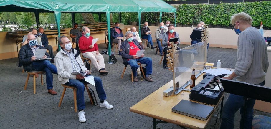 Die Mitglieder des Männerchors sitzen mit Masken auf Stühlen und proben im Freien mit viel Abstand.