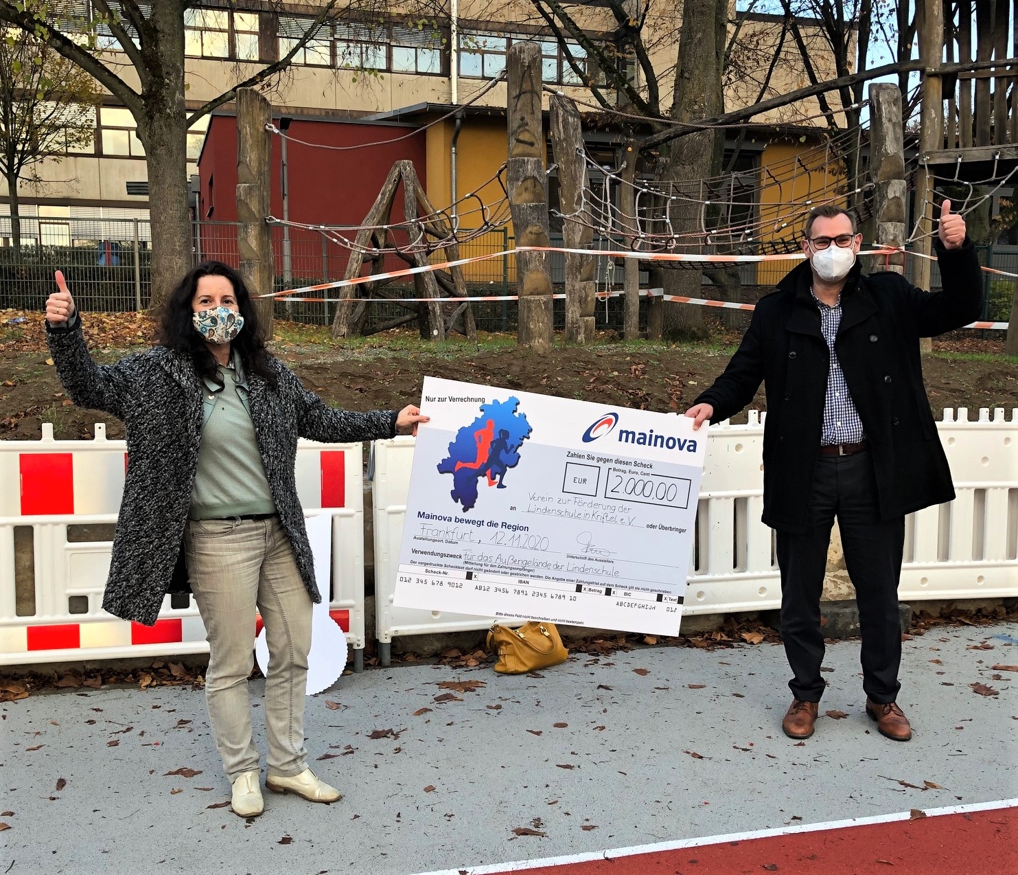 Bürgermeister Seitz und Ljudmilla Regeniter, Vorsitzende des Fördervereins Lindenschule, halten einen großen Scheck. Sie stehen auf dem Schulhof und jubeln.