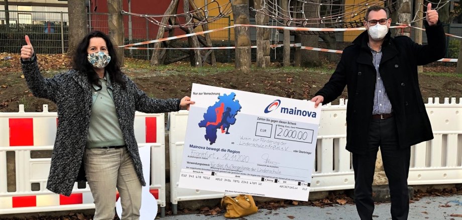 Bürgermeister Seitz und Ljudmilla Regeniter, Vorsitzende des Fördervereins Lindenschule, halten einen großen Scheck. Sie stehen auf dem Schulhof und jubeln.