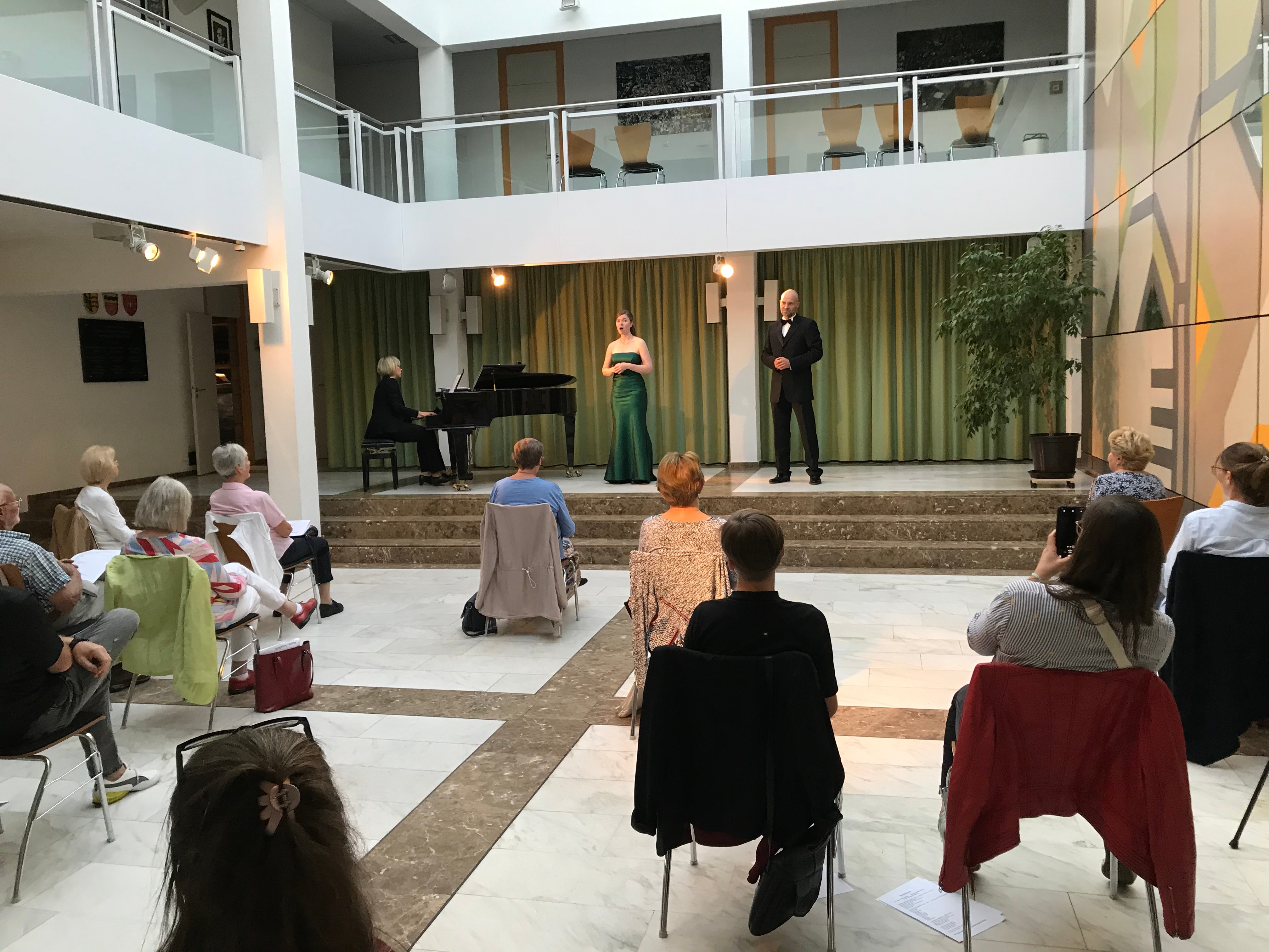 Zwei Sänger und eine Pianistin auf der Bühne, wenige Zuschauer auf weit entfernten Stühlen.