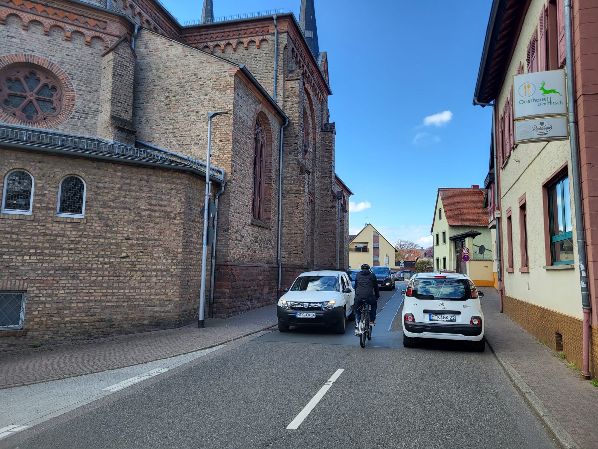 Ein Radfahrer wird neben einem parkenden Auto von einem entgegenkommenden Auto bedrängt.