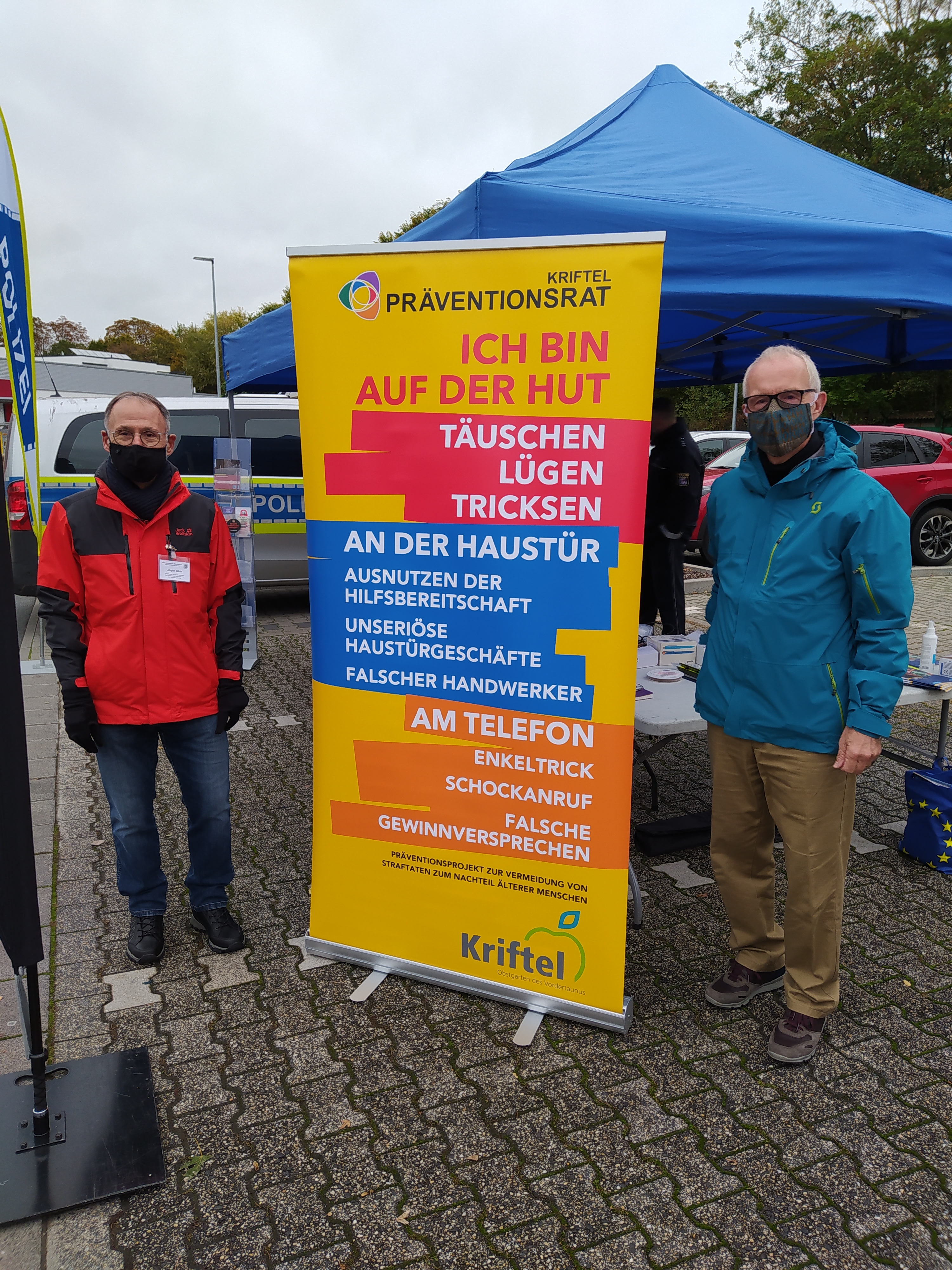 Zwei Sicherheitsberater mit Maske stehen neben einem großen Schild. Hier wird vor Trickbetrügern gewarnt.