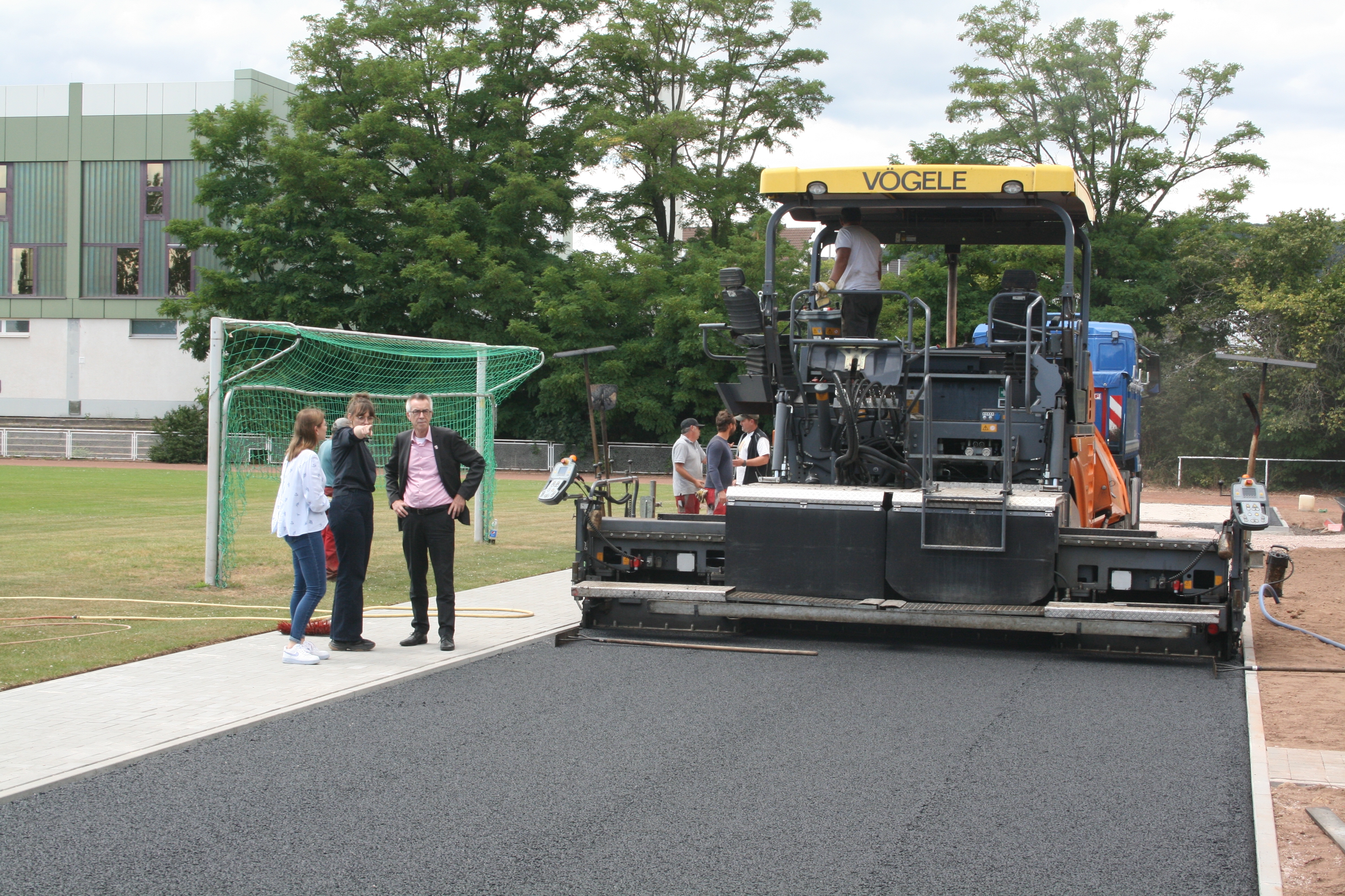 Eine riesige Baumaschine bringt Asphlat auf dem Sportplatz auf, da wo die Anlaufbahn der Weitspringer sein wird.