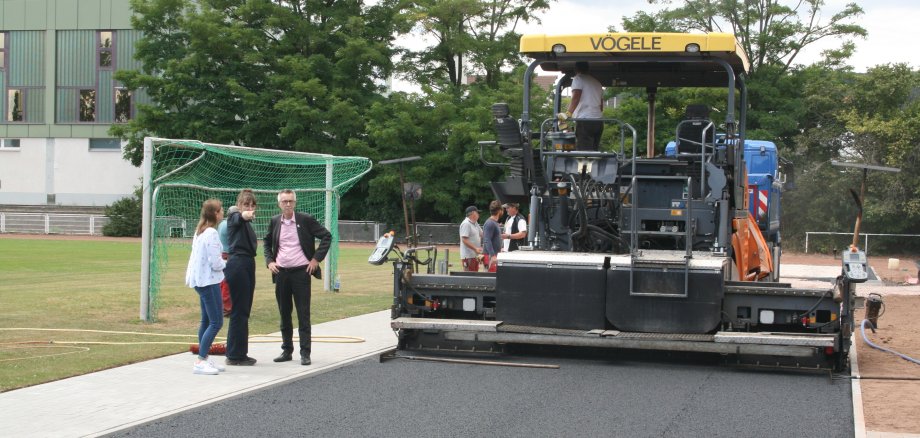 Eine riesige Baumaschine bringt Asphlat auf dem Sportplatz auf, da wo die Anlaufbahn der Weitspringer sein wird.