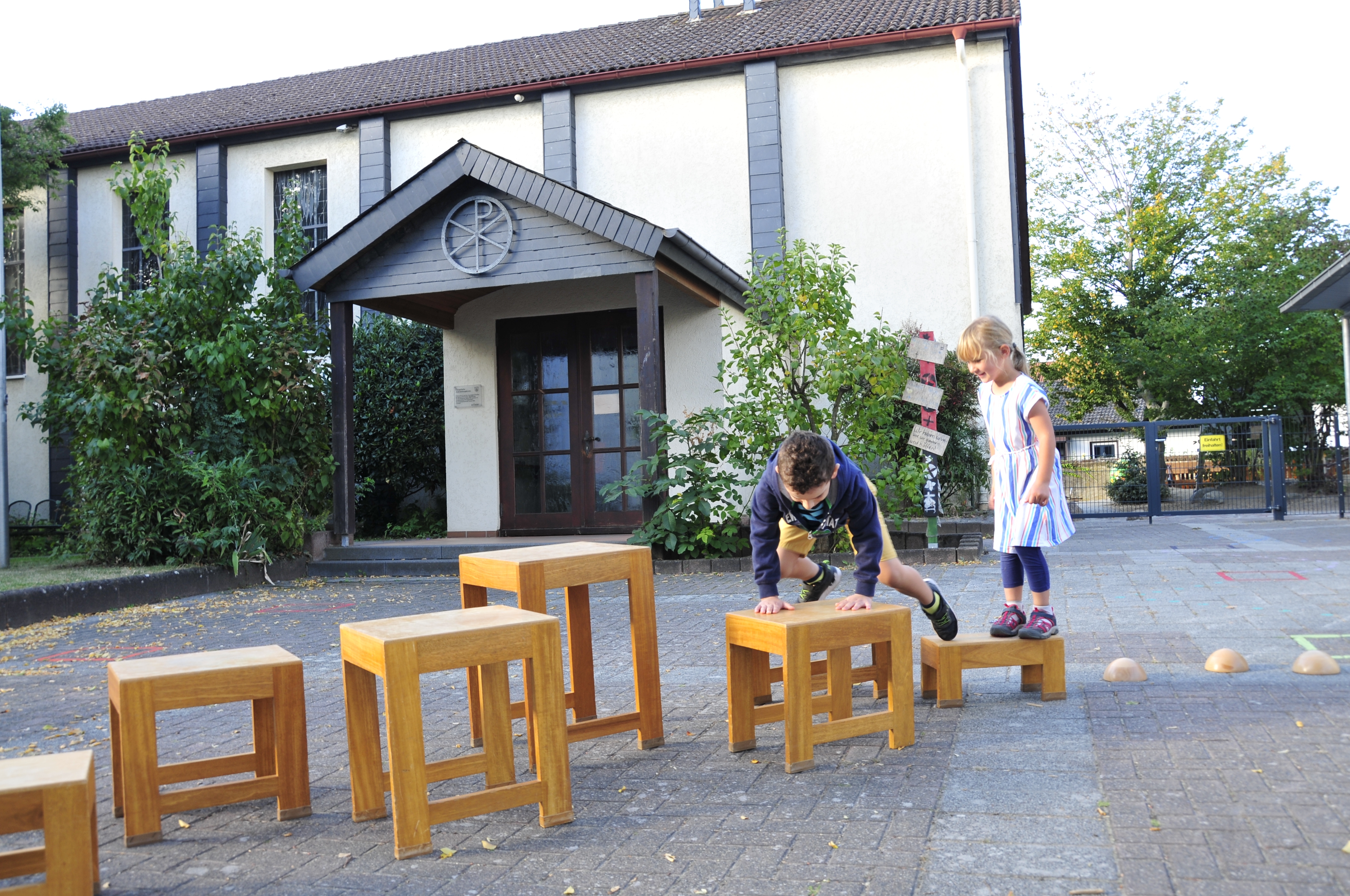 Kinder klettern vor der Kita über verschieden hohe Holzstühle.