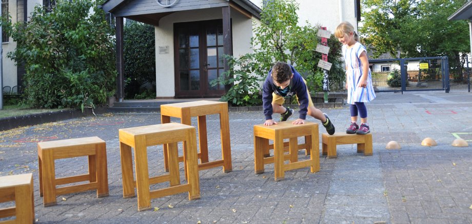 Kinder klettern vor der Kita über verschieden hohe Holzstühle.
