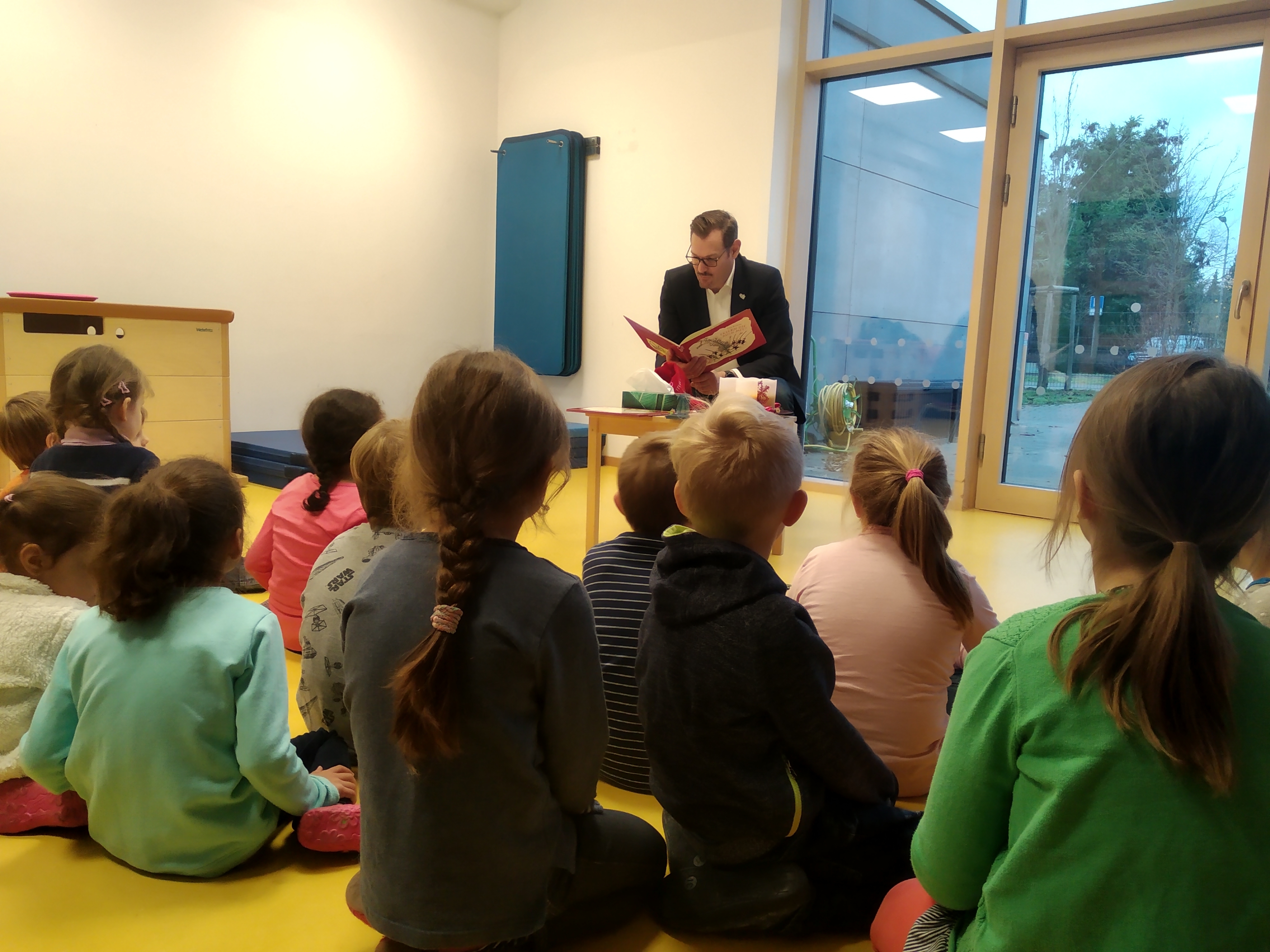 Um den Bürgermeister mit Buch in der Hand sitzen viele Kinder herum.