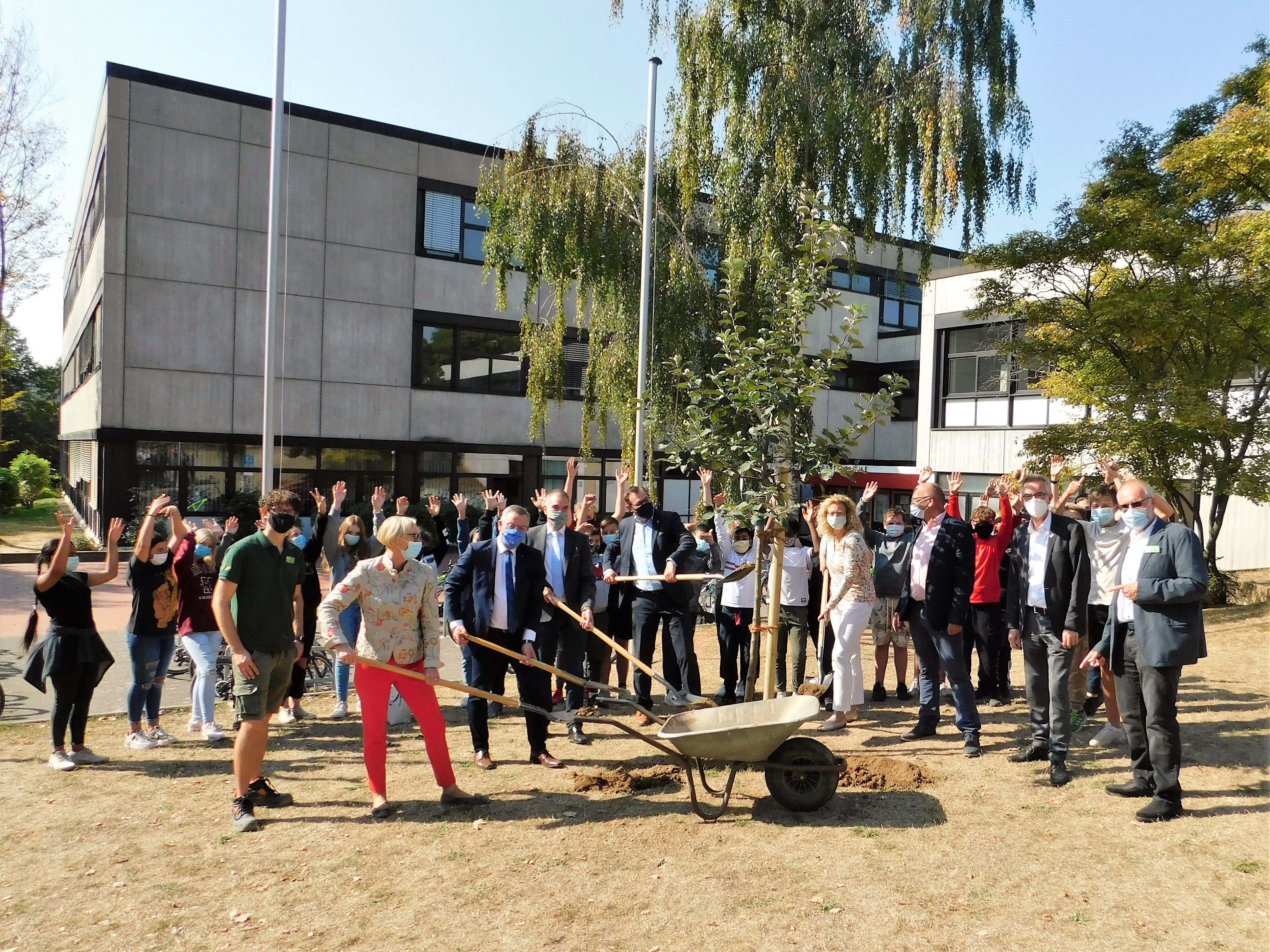 Viel Prominenz mit Maske und Schaufel pflanzt einen Baum vor die Weingartenschule.